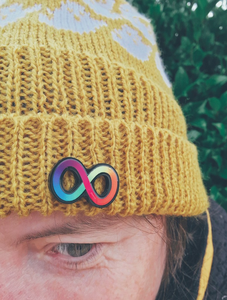 Close up of a woman in a yellow knitted hat with white ghosts on it. On the brim there is a Neuro pride pin badge (the infinity symbol in rainbow colours).